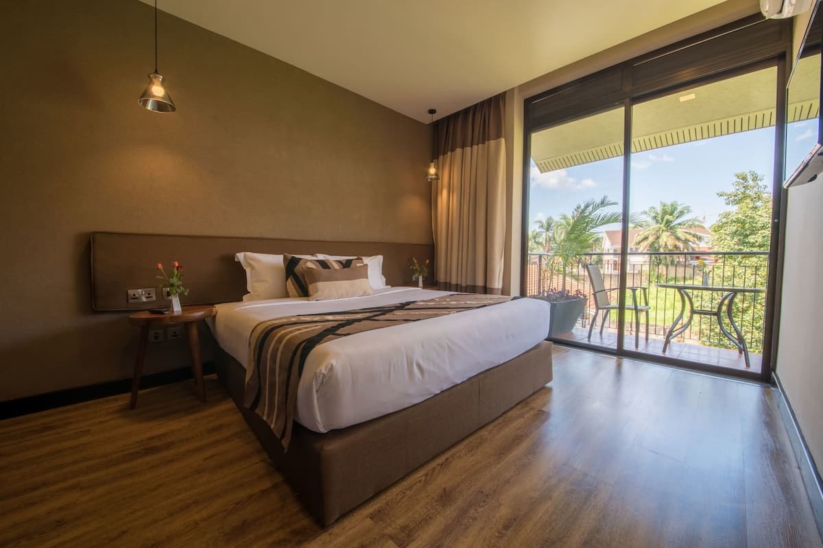 A hotel room with a large bed, a nightstand, a small table, and a balcony with a view of palm trees.