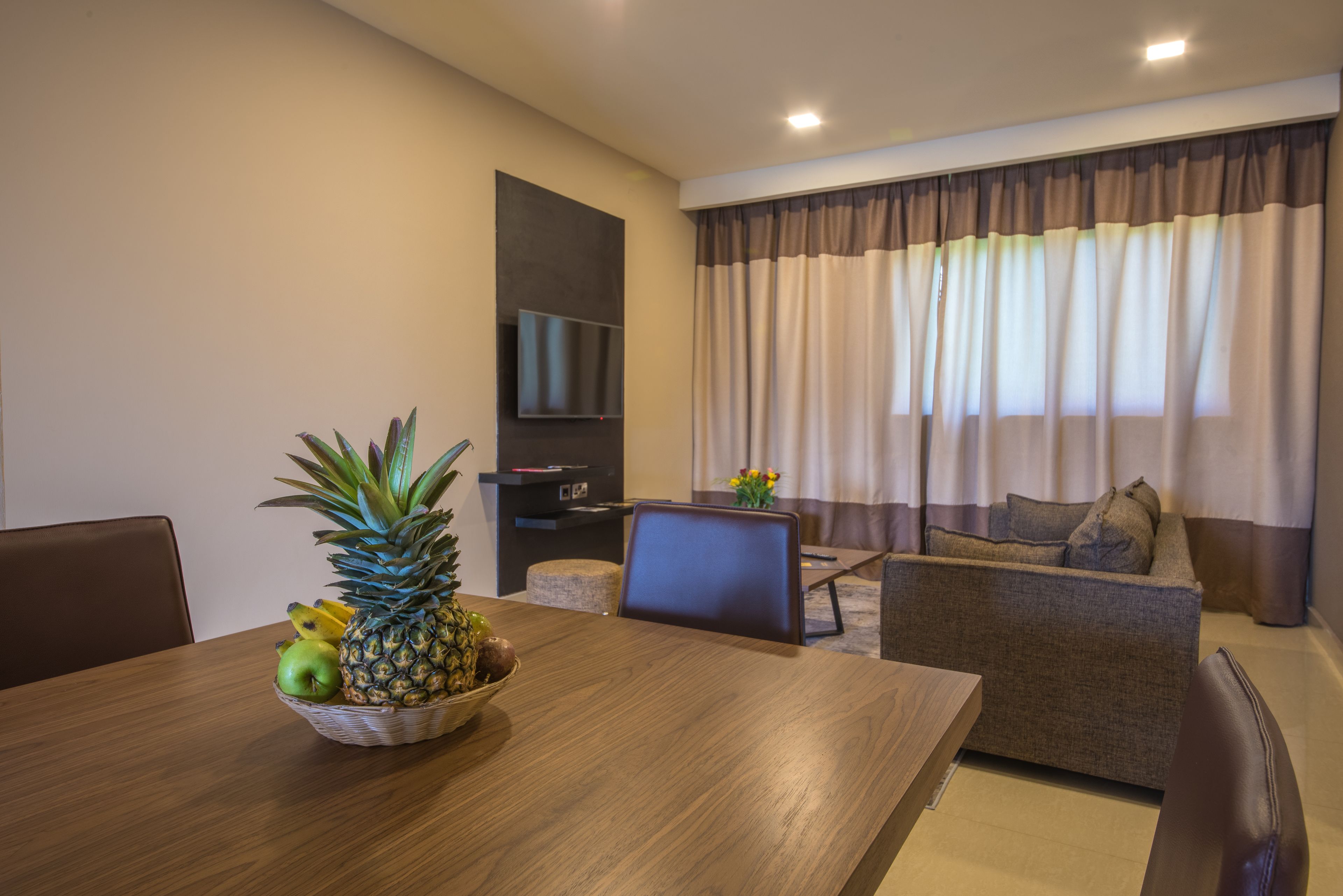 A modern living room with a wooden dining table, a sofa, and a TV mounted on the wall.