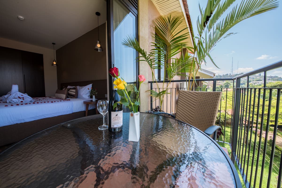 A room with a bed, a table with a bottle and glasses, and a balcony with plants.