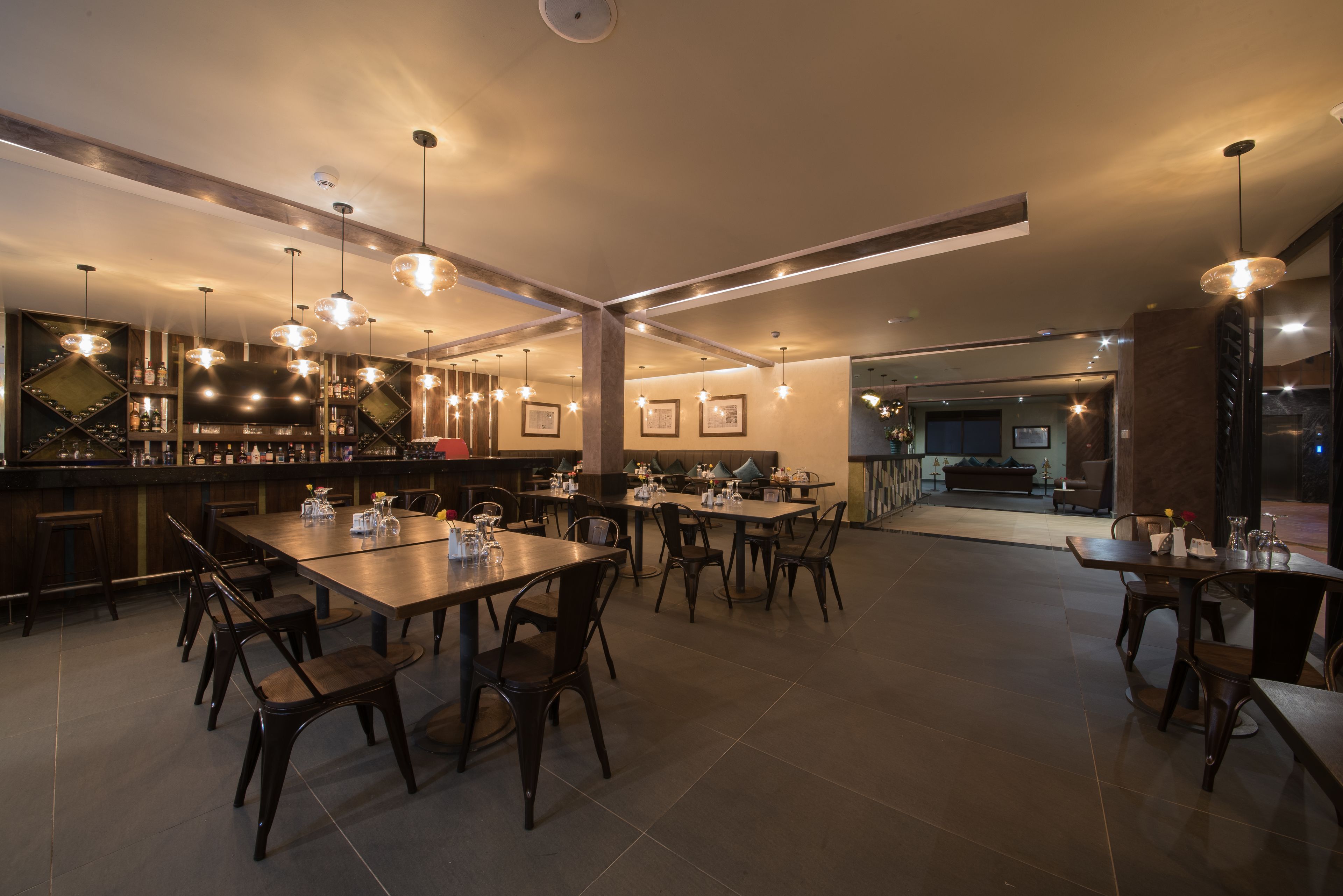 A modern restaurant with multiple dining tables and chairs, a bar area with shelves of bottles, and pendant lighting.