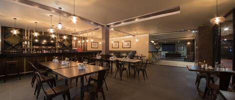 A modern restaurant with multiple dining tables and chairs, a bar area with shelves of bottles, and pendant lighting.
