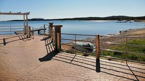 A paved walkway leading to a body of water with a metal railing and a pergola structure. - Aria di Vacanza- Molara (Loiri Porto San Paolo)