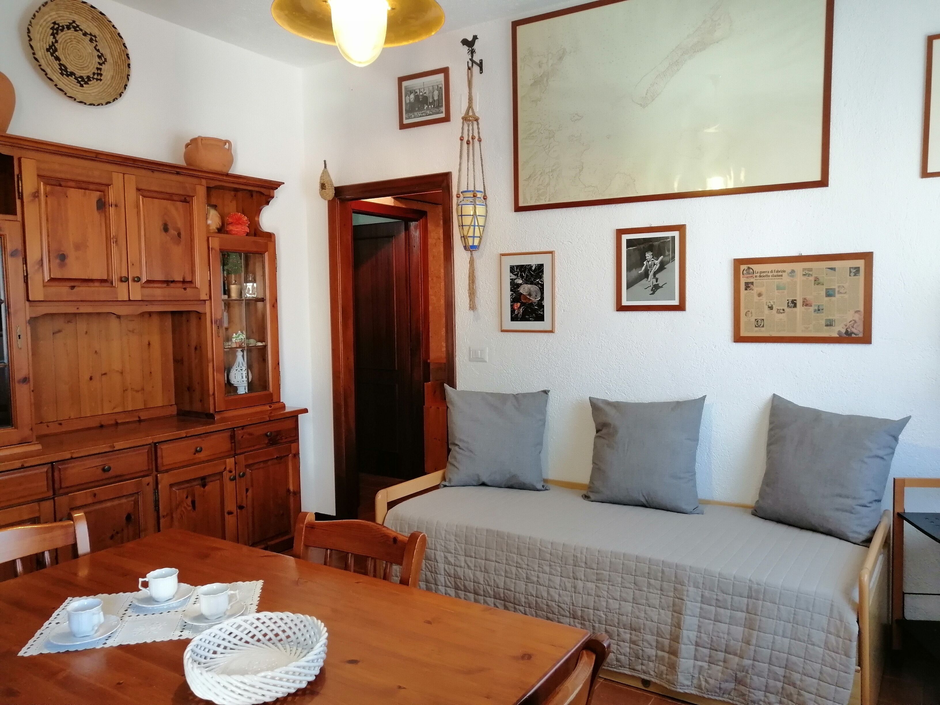 A cozy room with a wooden dining table set for four, a sofa with cushions, a large framed picture, and a hanging light fixture.