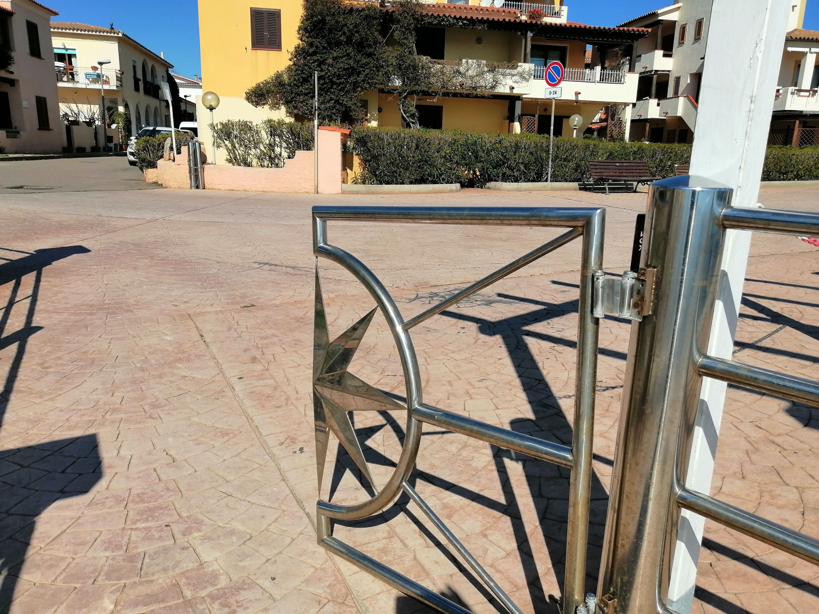 A gated entrance with a metal gate, a paved area, and buildings with balconies in the background.