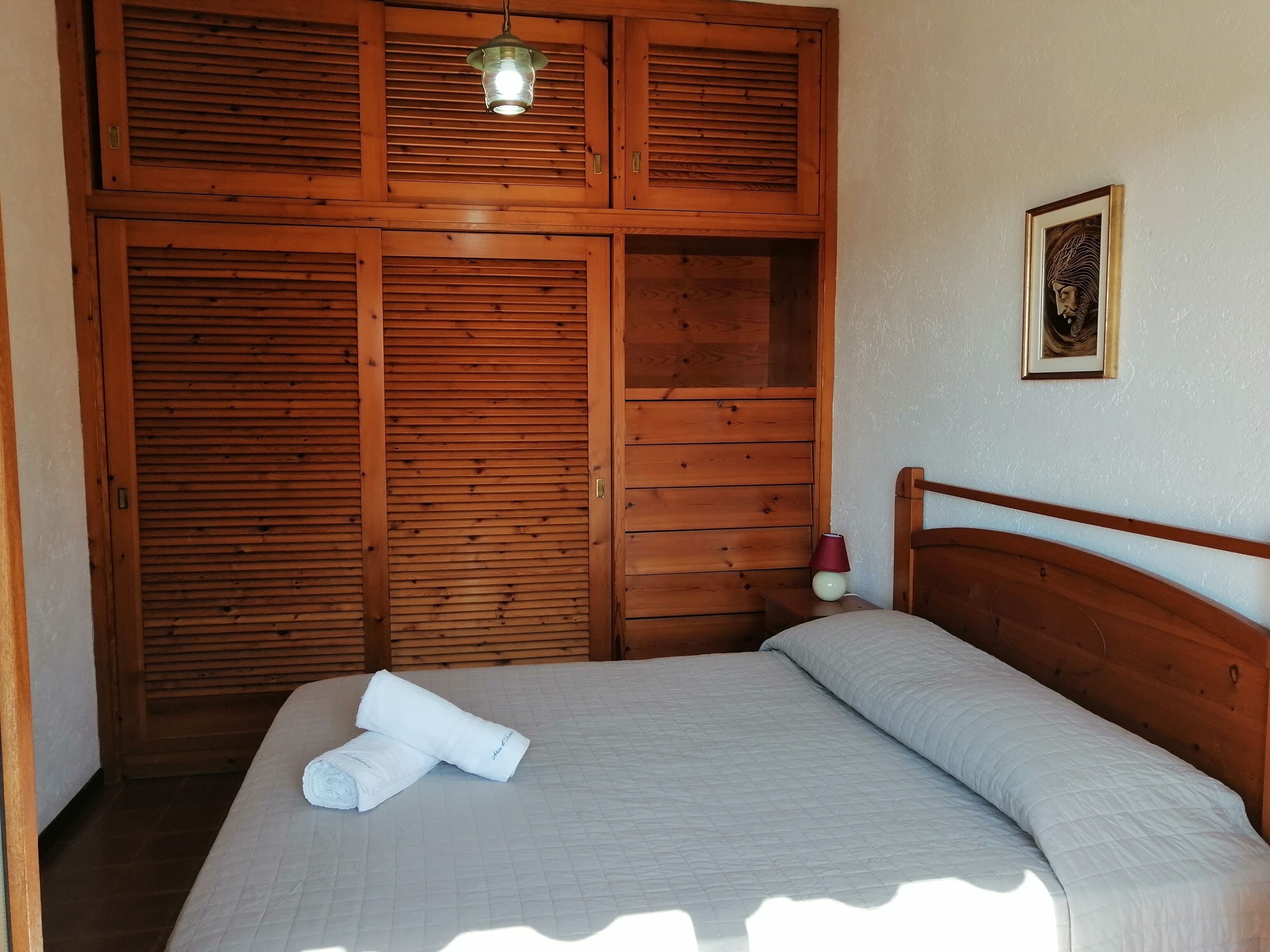 A bedroom with a wooden wardrobe, a bed with a white duvet, and a small bedside table with a lamp.