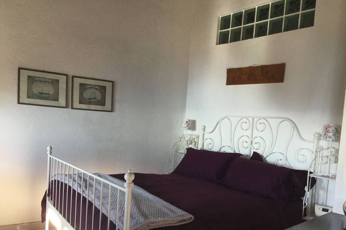 Un dormitorio con cama, mesita de noche, ventana y dos cuadros en la pared.