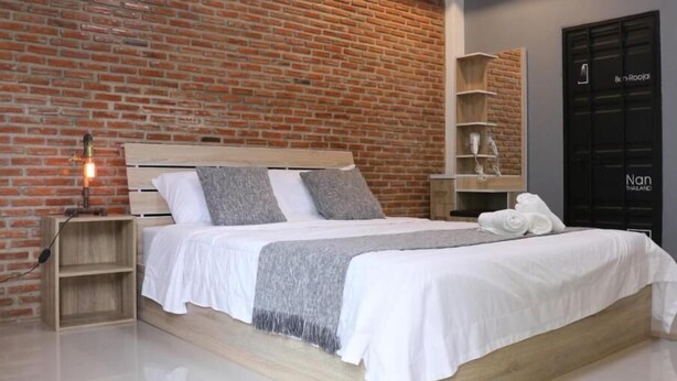 A modern bedroom with a brick wall, a wooden bed frame, white bedding, and a bedside lamp. - Ban Roojai (Nan)