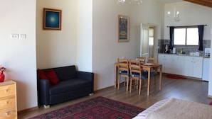 A living room with a dark blue sofa, a wooden dining table with chairs, a kitchen area with white cabinets, and a chandelier. - Off The Square (Safed)