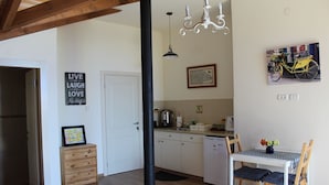 A compact kitchen with a dining area, wooden cabinets, a black column, a chandelier, and a bicycle painting on the wall. - Off The Square (Safed)