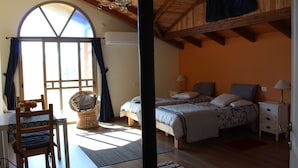A bedroom with a large window, wooden ceiling beams, a bed with two pillows, a nightstand, and a wicker chair. - Off The Square (Safed)