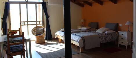 Un dormitorio con un ventanal grande, vigas de madera en el techo, una cama con dos almohadas, una mesita de noche y una silla de mimbre.