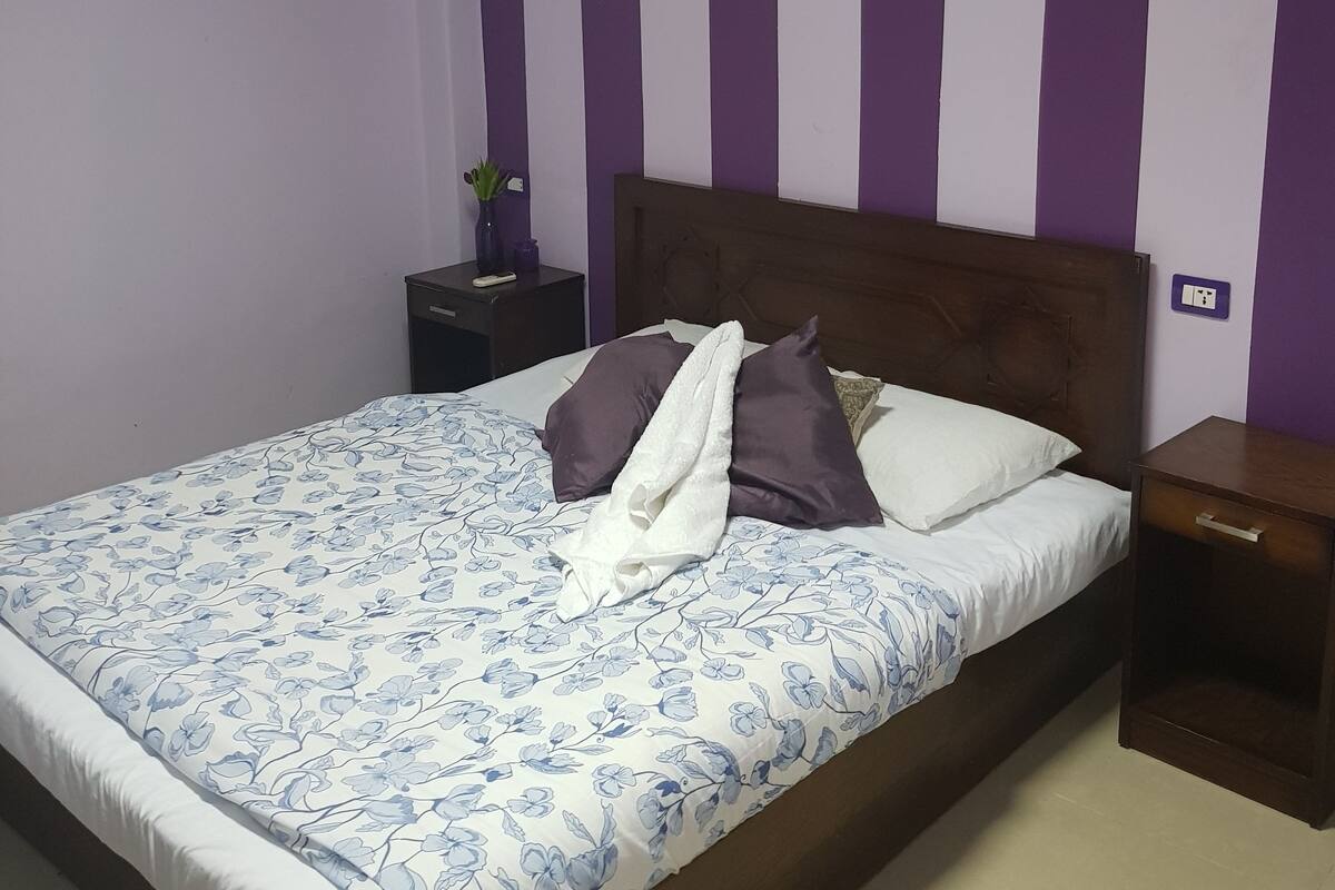 A neatly made bed with a floral-patterned bedspread, a wooden headboard, and a nightstand with a lamp and a small plant.