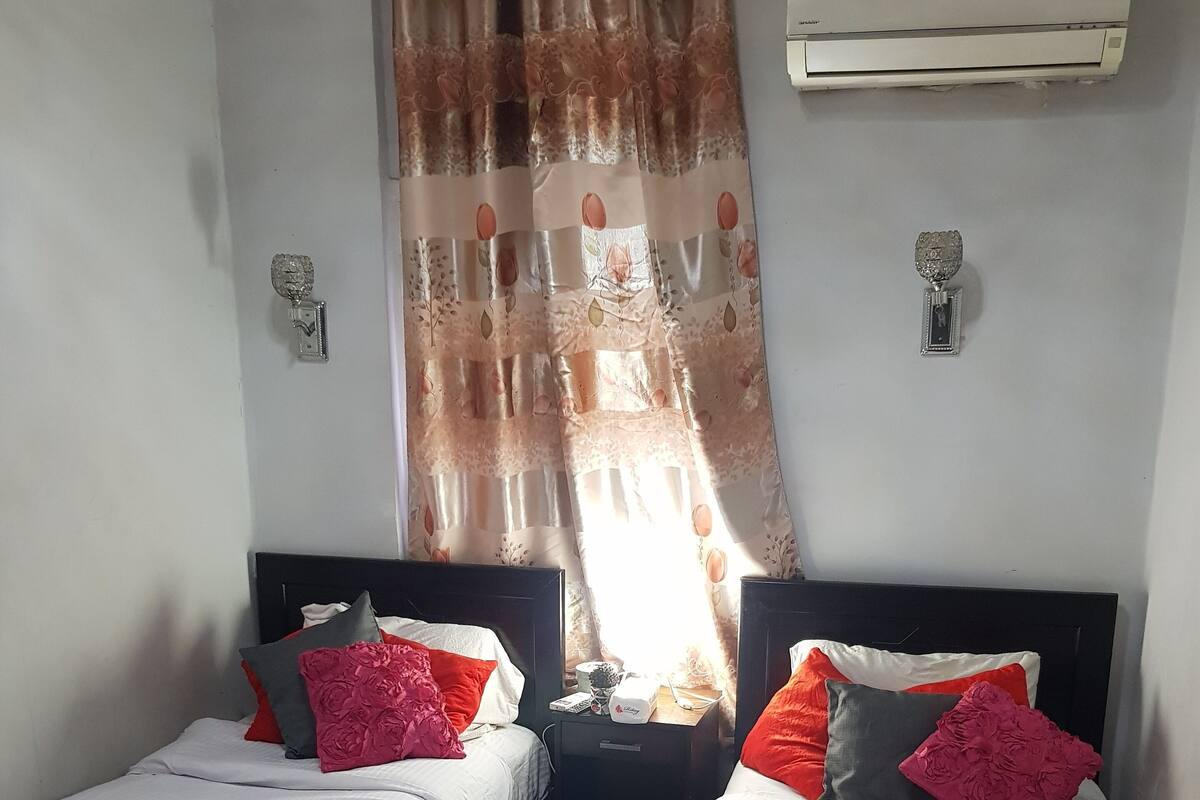 A room with two single beds, each with a white duvet and red pillows. There is a nightstand between the beds with a small table and a lamp. A window with a floral curtain is visible, and an air conditioning unit is mounted on the wall.