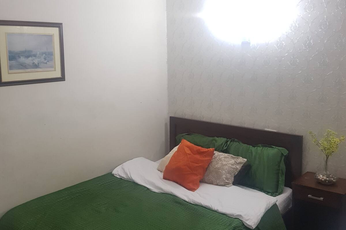 A neatly made bed with green bedding and multiple pillows, a small bedside table with a vase, and a framed picture on the wall.