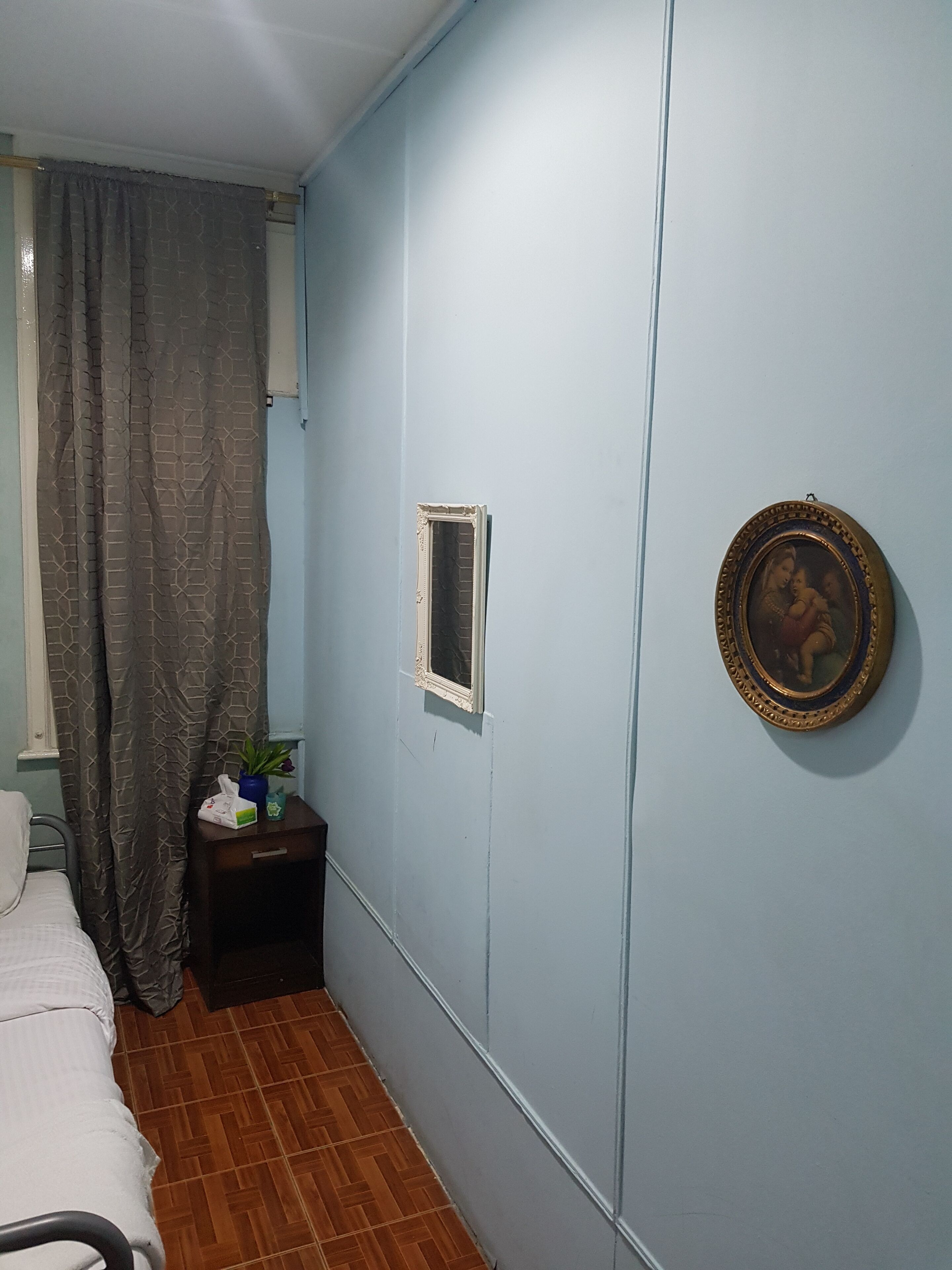 A small, narrow room with a bed, a nightstand, a curtain, a framed picture on the wall, and a window with a curtain.