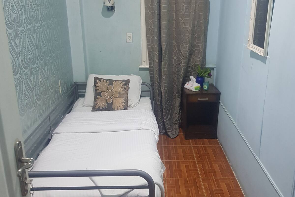 A small, neatly arranged bedroom with a single bed, a nightstand, and a window with a curtain.