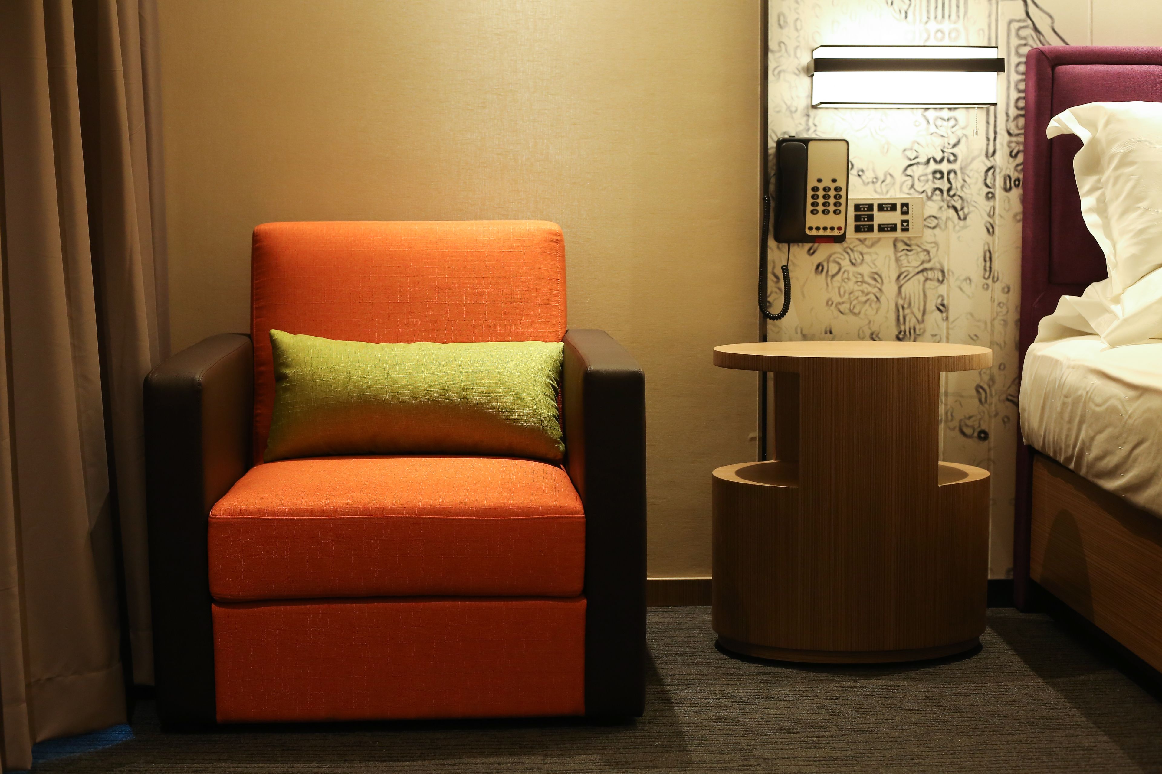 Habitación de hotel con una sola silla naranja, una pequeña mesita de madera, un teléfono y una cama con ropa blanca.