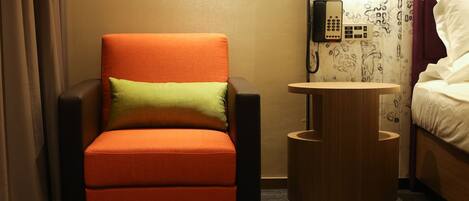 Habitación de hotel con una sola silla naranja, una pequeña mesita de madera, un teléfono y una cama con ropa blanca.