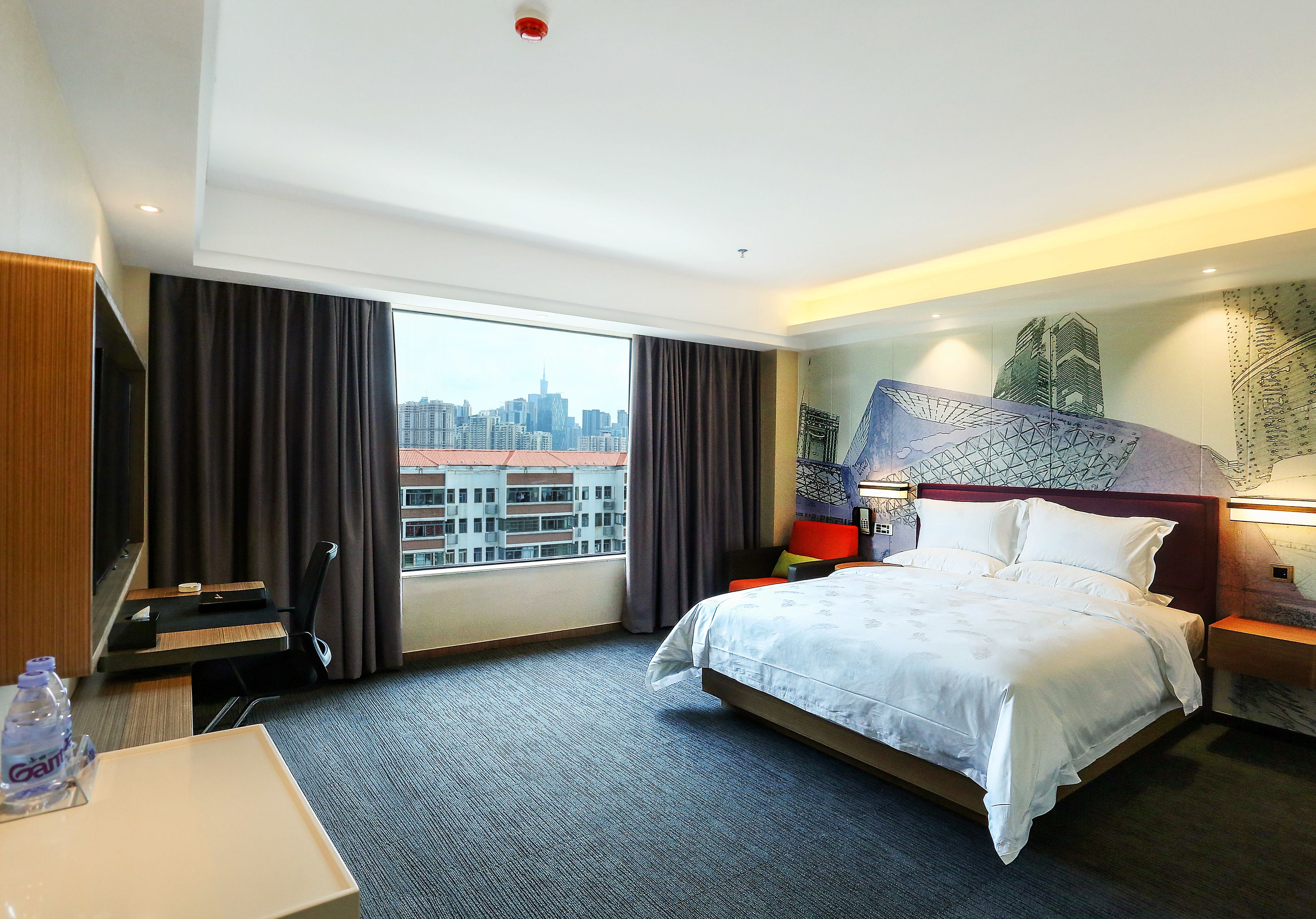 A hotel room with a large bed, a desk, a chair, a TV, and a view of the cityscape through the window.