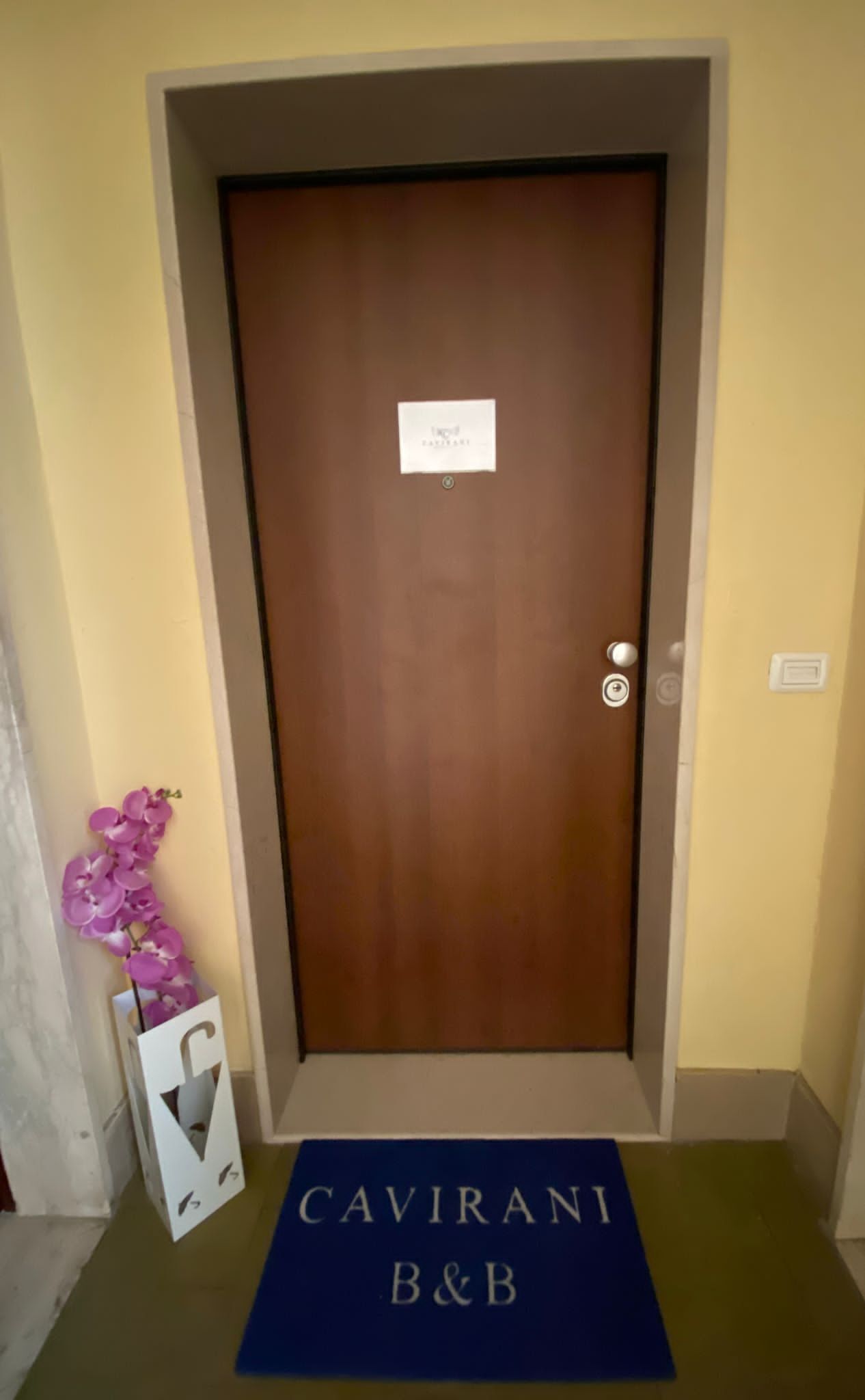 A wooden door with a sign above it, a purple orchid in a white box, and a blue mat with "CAVIRANI B&B" on it.
