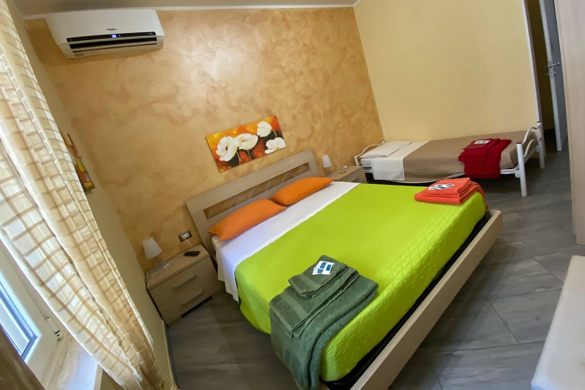 A hotel room with a bed, a nightstand, a bedside table, an air conditioner, a painting, and a window with curtains.