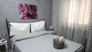 A bedroom with a bed, a nightstand, a lamp, a wall with a floral painting, and a window with curtains. - B&B Cavirani (Gallipoli)