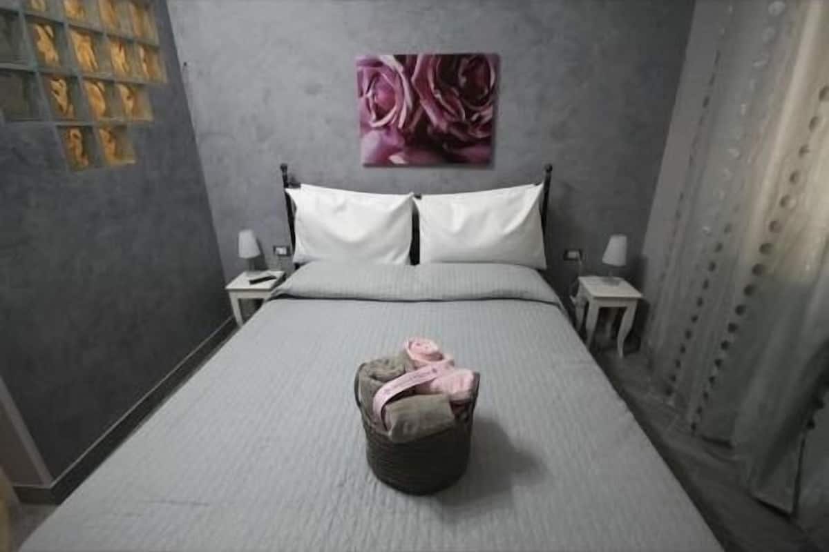 A bedroom with a bed, pillows, a bedside table, a lamp, a wall with a floral painting, and a curtain.