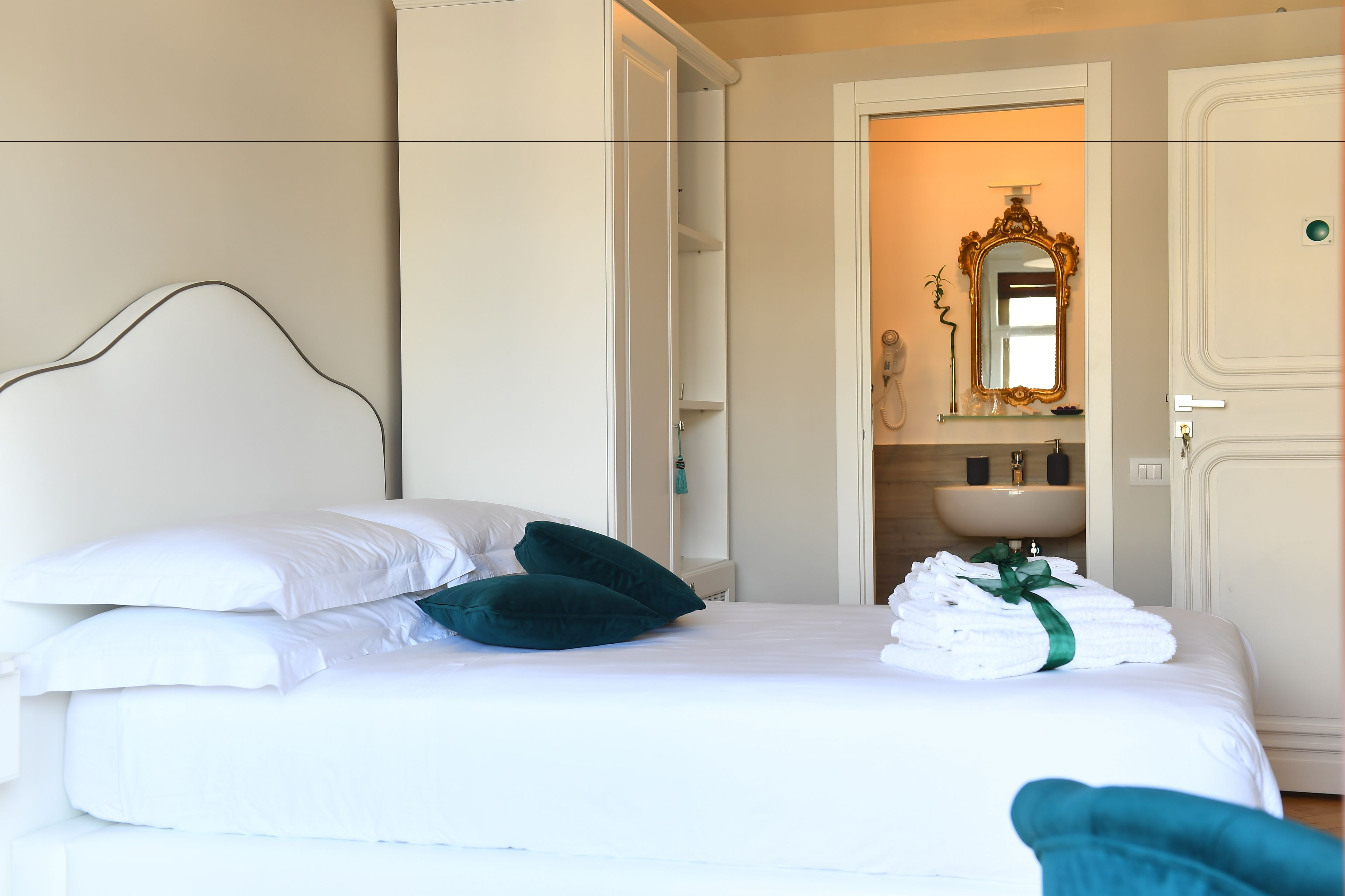 A neatly made bed with white linens and teal pillows, a bathroom with a mirror and sink, and a door leading to another room.