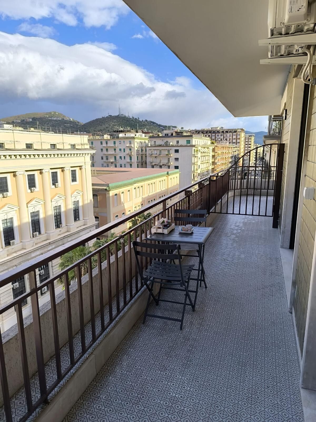 Un balcone con tavolo e sedie, affaccia su un panorama urbano con edifici e una collina.
