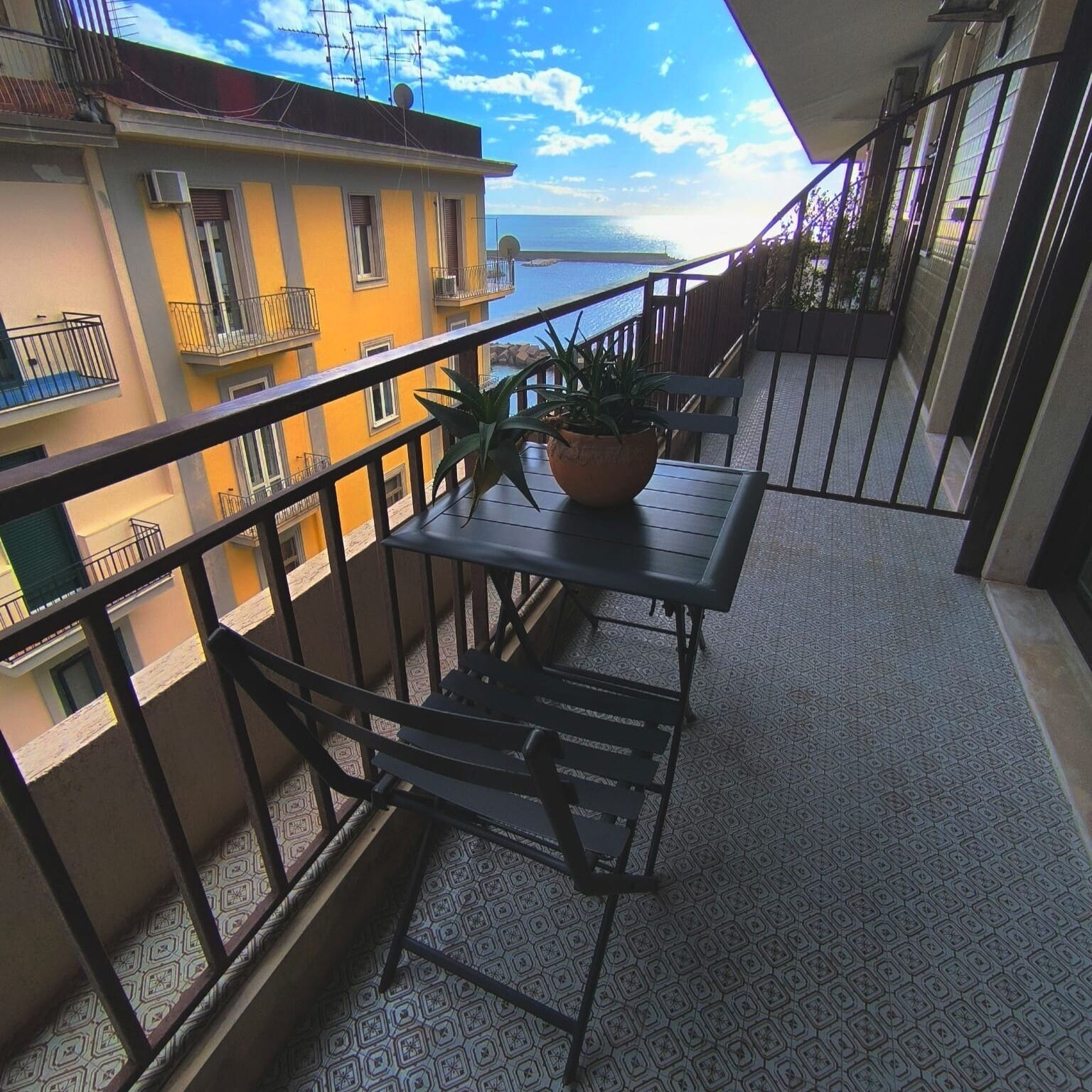 A balcony with a table and chairs, a potted plant, and a view of the sea.