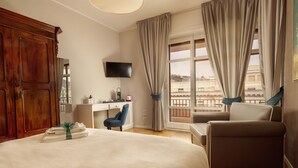 Quarto de hotel com uma cama grande, uma escrivaninha com um computador, uma cadeira e um sofá. Há uma televisão na parede e uma janela com cortinas.