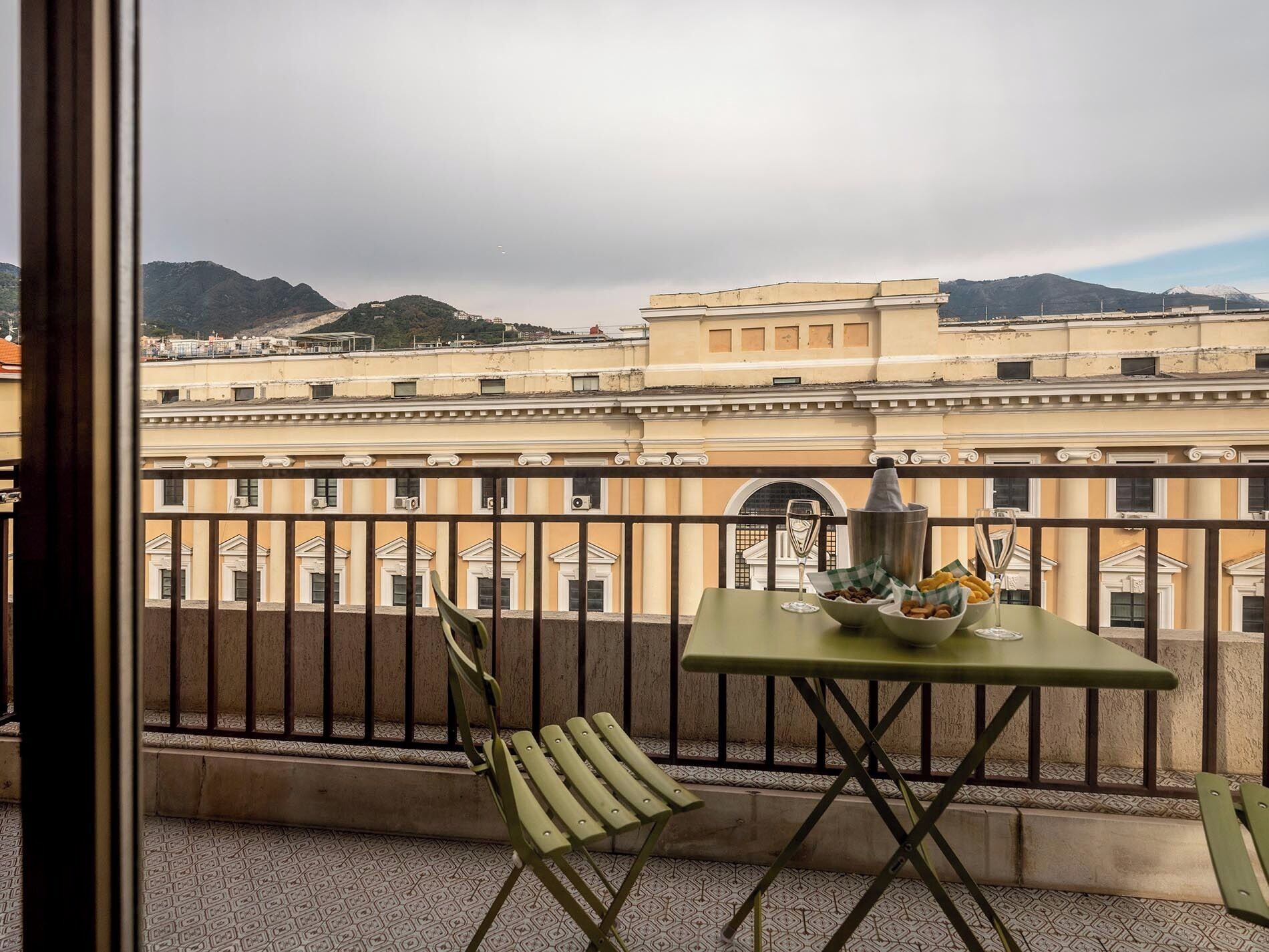 A balcony with a table set for two, a chair, and a view of a historic building with columns and a mountain in the background.
