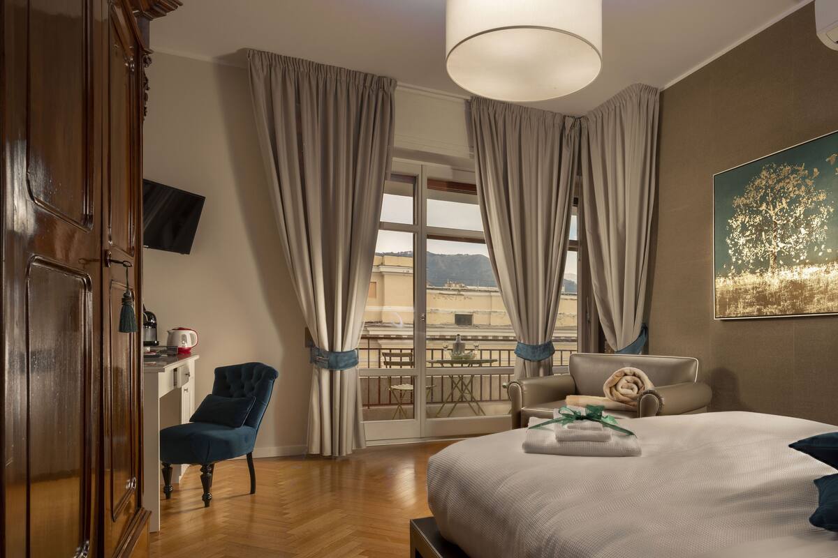 A hotel room with a large bed, a wooden wardrobe, a blue armchair, a small table with a kettle, and a balcony with a view of the city.