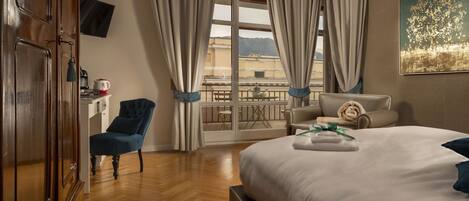 A hotel room with a large bed, a wooden wardrobe, a blue armchair, a small table with a kettle, and a balcony with a view of the city.