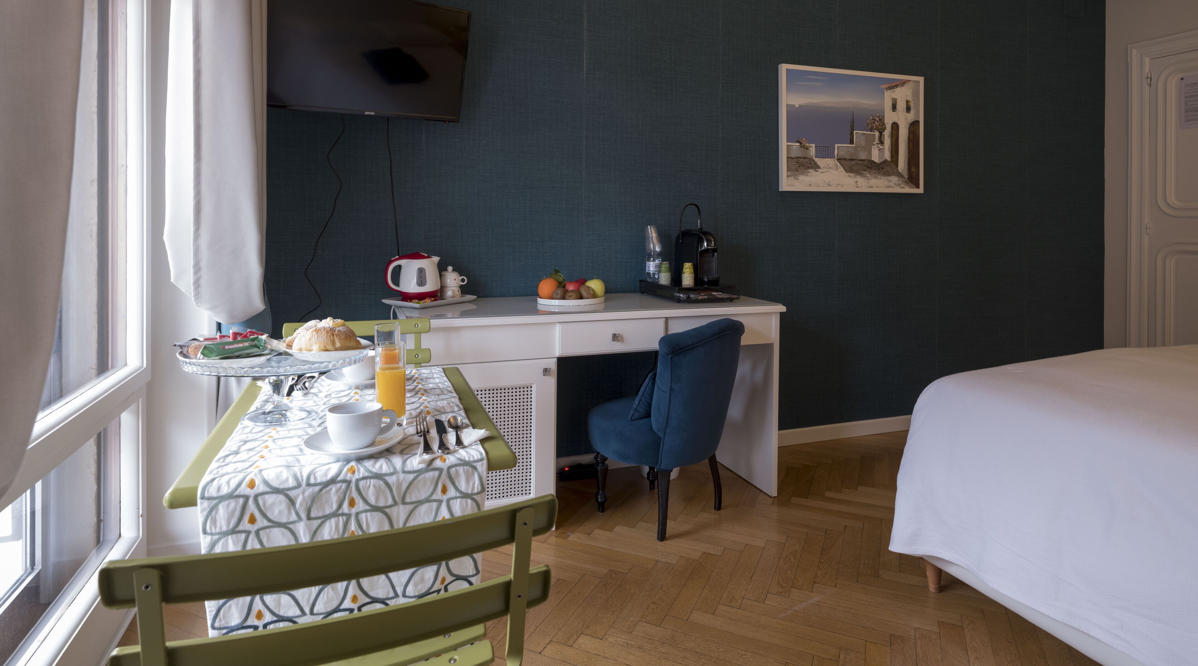 A hotel room with a bed, a desk with breakfast items, a chair, and a painting on the wall.