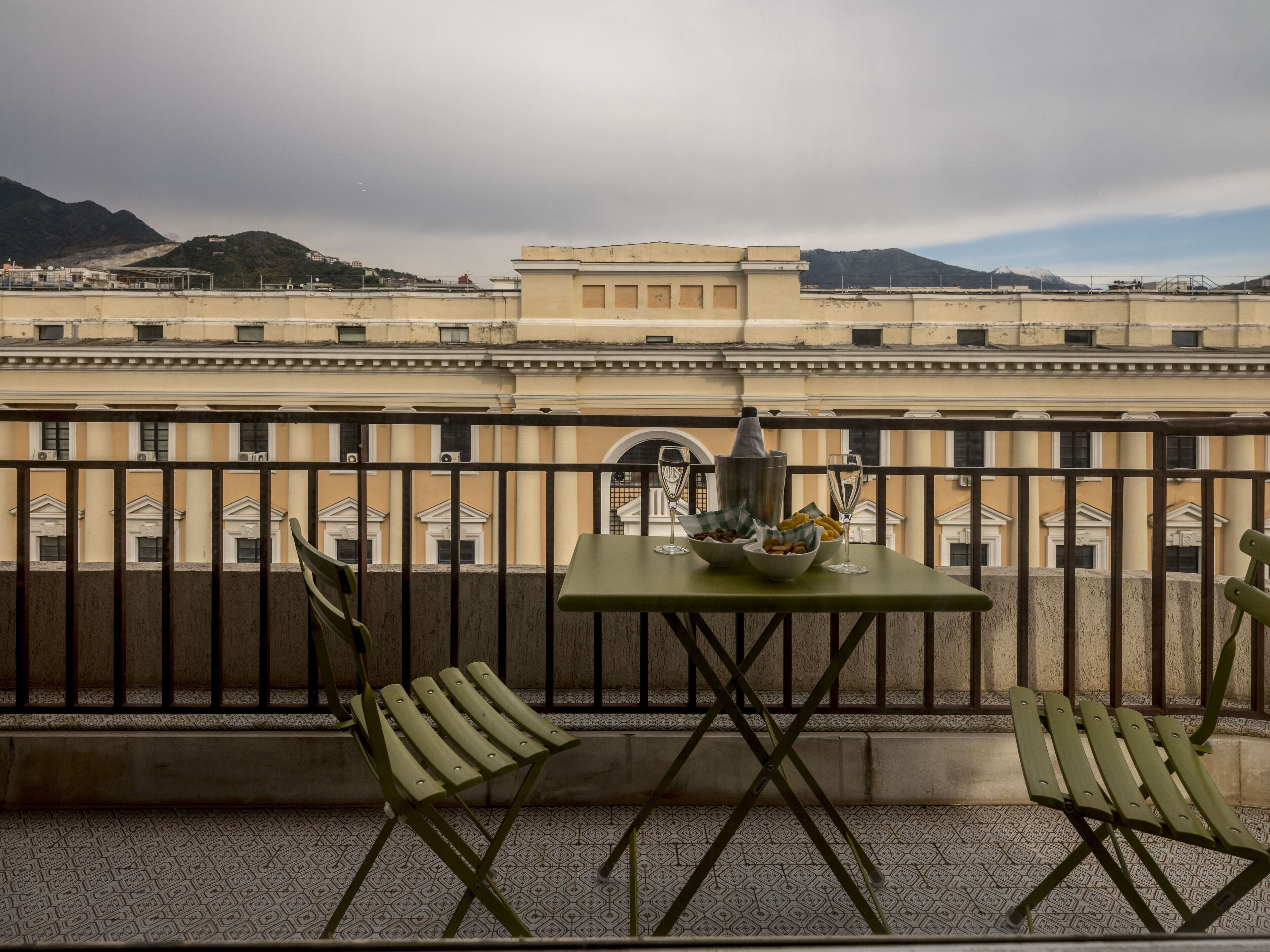 Un balcon avec une table dressée pour deux personnes, une bouteille et un verre. Le balcon offre une vue sur un immeuble à l’architecture classique, avec des montagnes en arrière-plan.