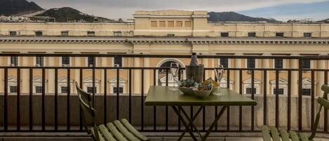 Un balcon avec une table dressée pour deux personnes, une bouteille et un verre. Le balcon offre une vue sur un immeuble à l’architecture classique, avec des montagnes en arrière-plan.