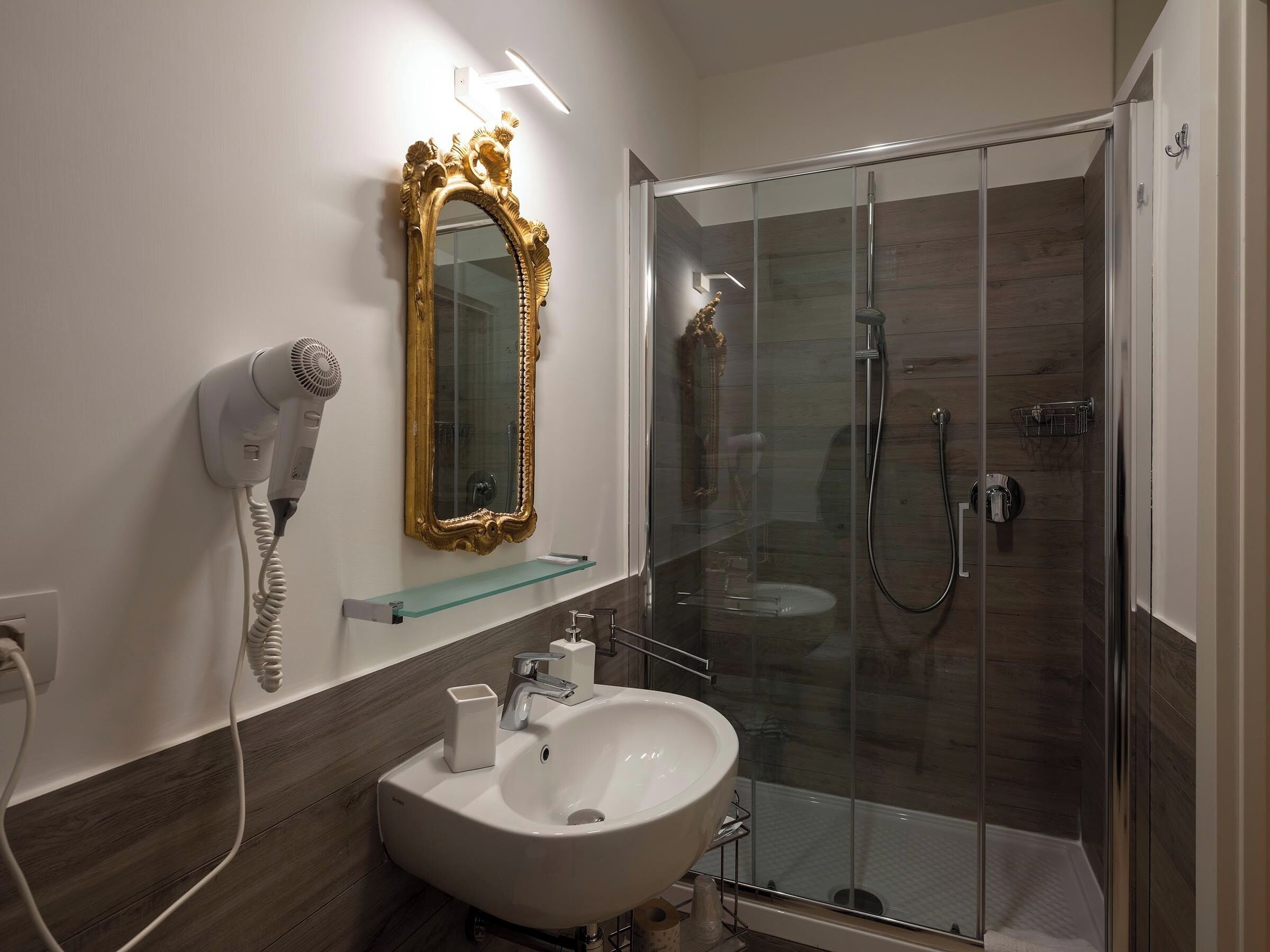 A bathroom with a white sink, a gold-framed mirror, a hairdryer, and a glass-enclosed shower.