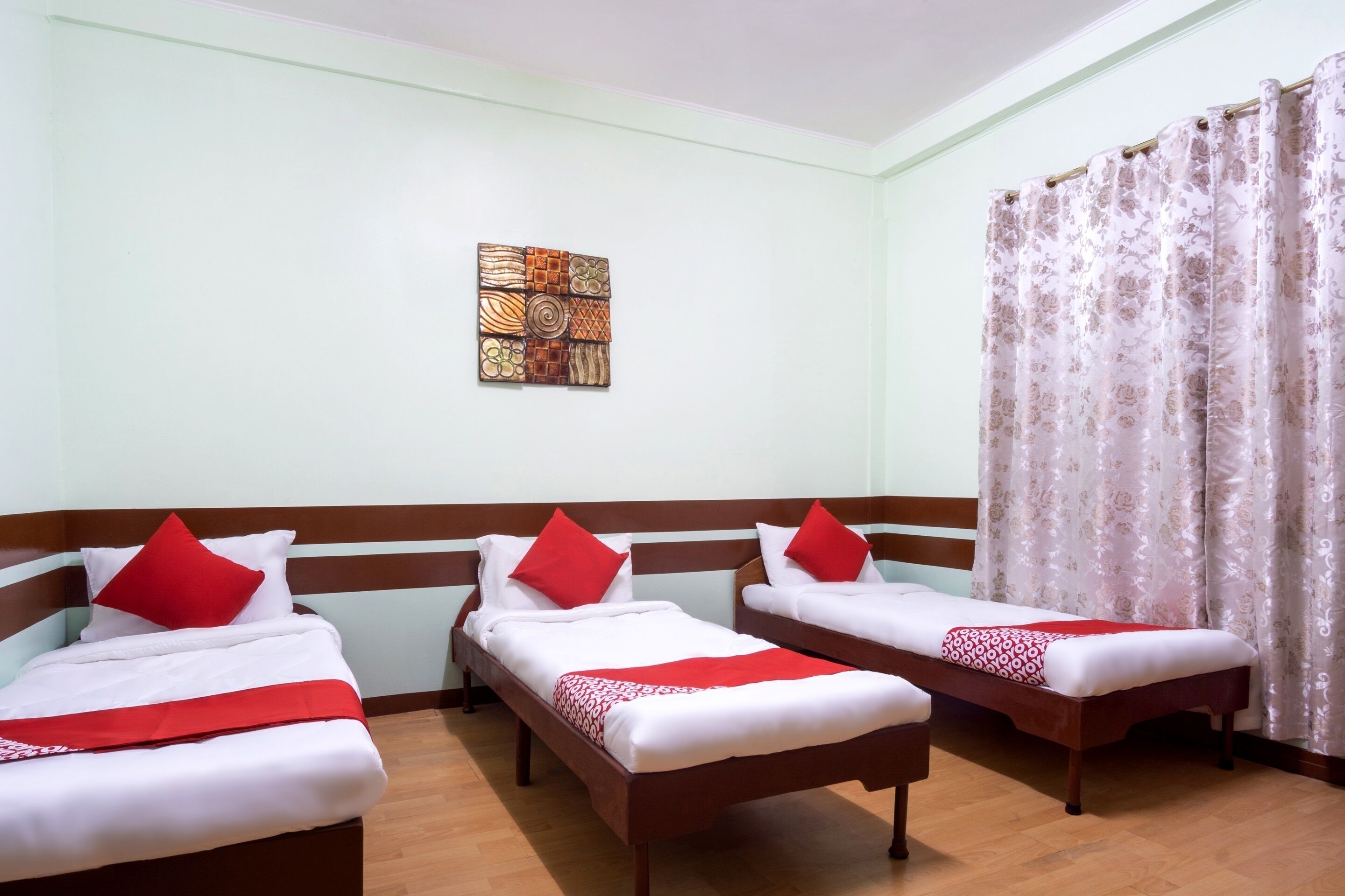 A hotel room with three single beds, each with a red pillow and a white bedspread. There is a wooden headboard, a small artwork on the wall, and a window with a floral curtain.