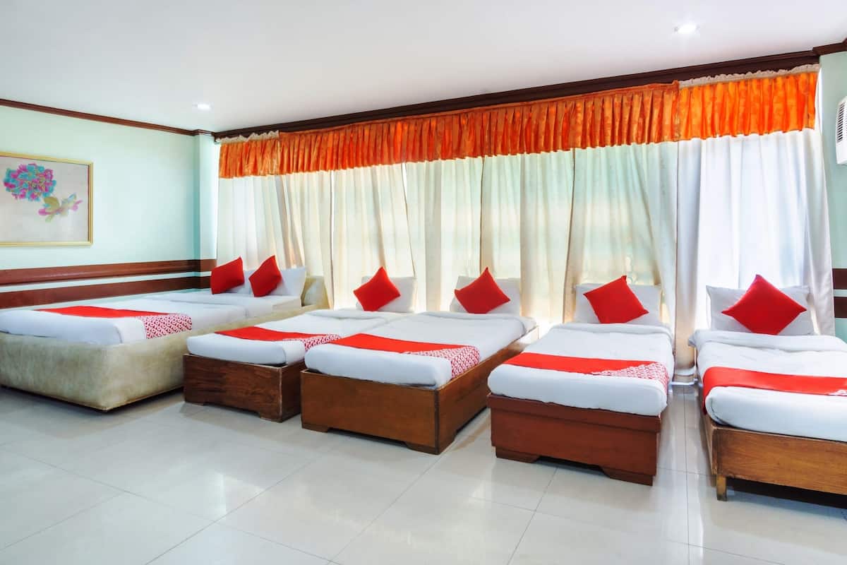 Une chambre d’hôtel avec quatre lits alignés, chacun étant doté de draps blancs et d’oreillers rouges. La chambre dispose d’une grande fenêtre avec des rideaux orange, d’un ventilateur de plafond et d’un climatiseur fixé au mur.