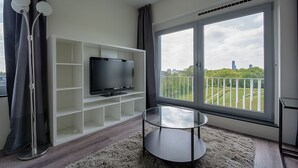 A modern living room with a flat-screen TV on a white shelving unit, a glass coffee table, and a large window with a view of a cityscape and greenery. - StudentStay (Leeuwarden)
