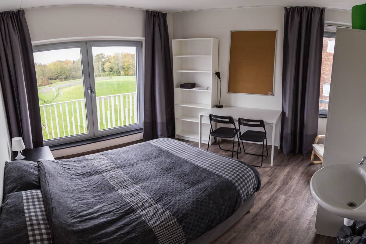 A bedroom with a large bed, a desk, two chairs, a whiteboard, and a view of a green landscape through a window.