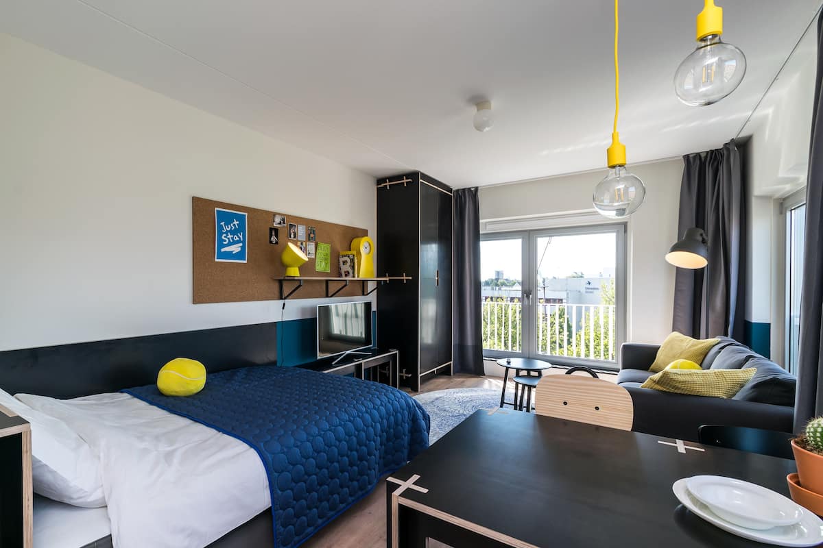 A modern hotel room with a bed, a desk, a television, a corkboard with photos, and a balcony with a view.