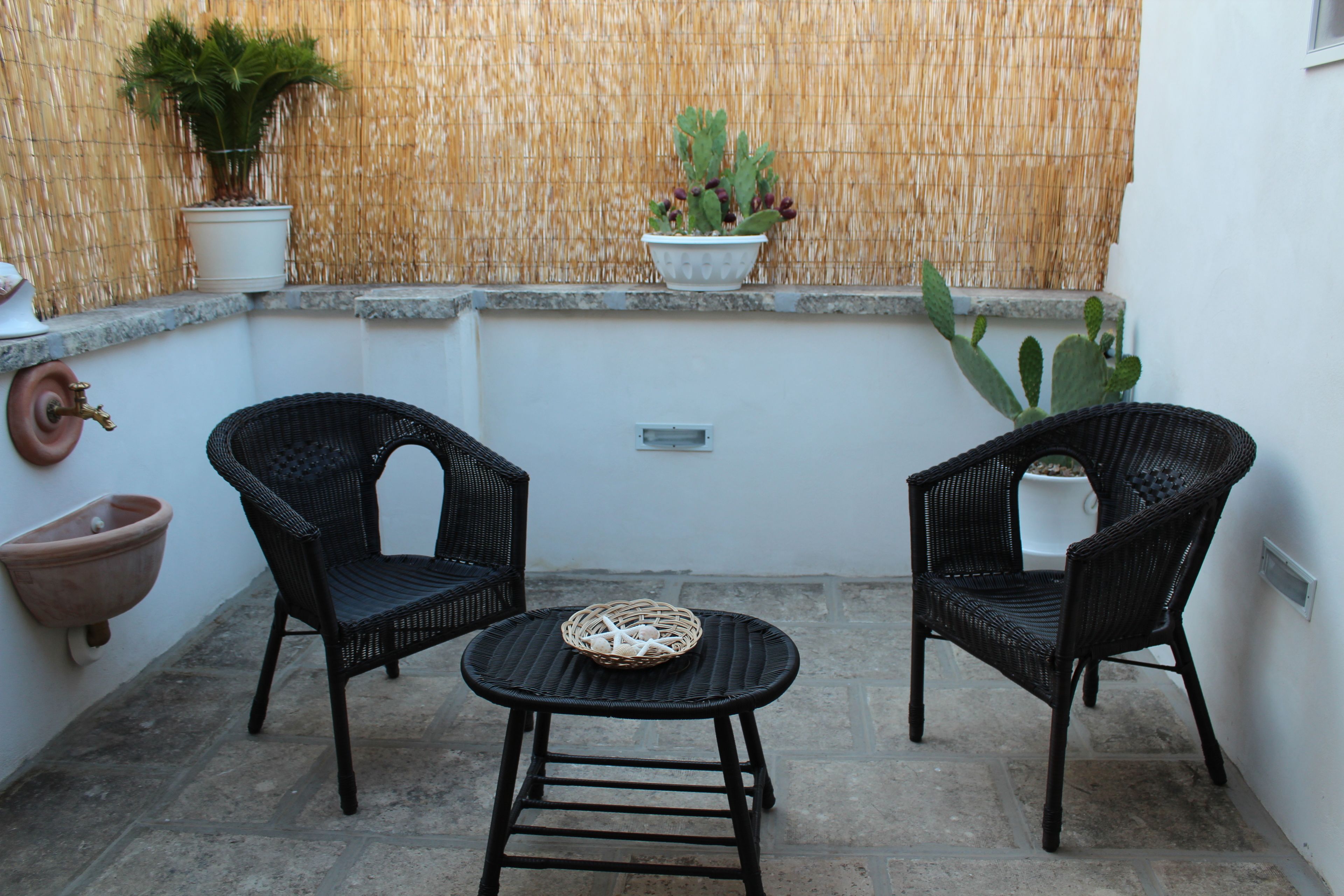 A small outdoor seating area with two wicker chairs and a round table, surrounded by potted plants and a bamboo wall.