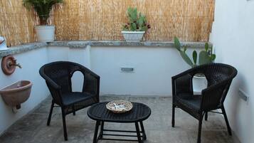 A small outdoor seating area with two wicker chairs and a round table, surrounded by potted plants and a bamboo wall.