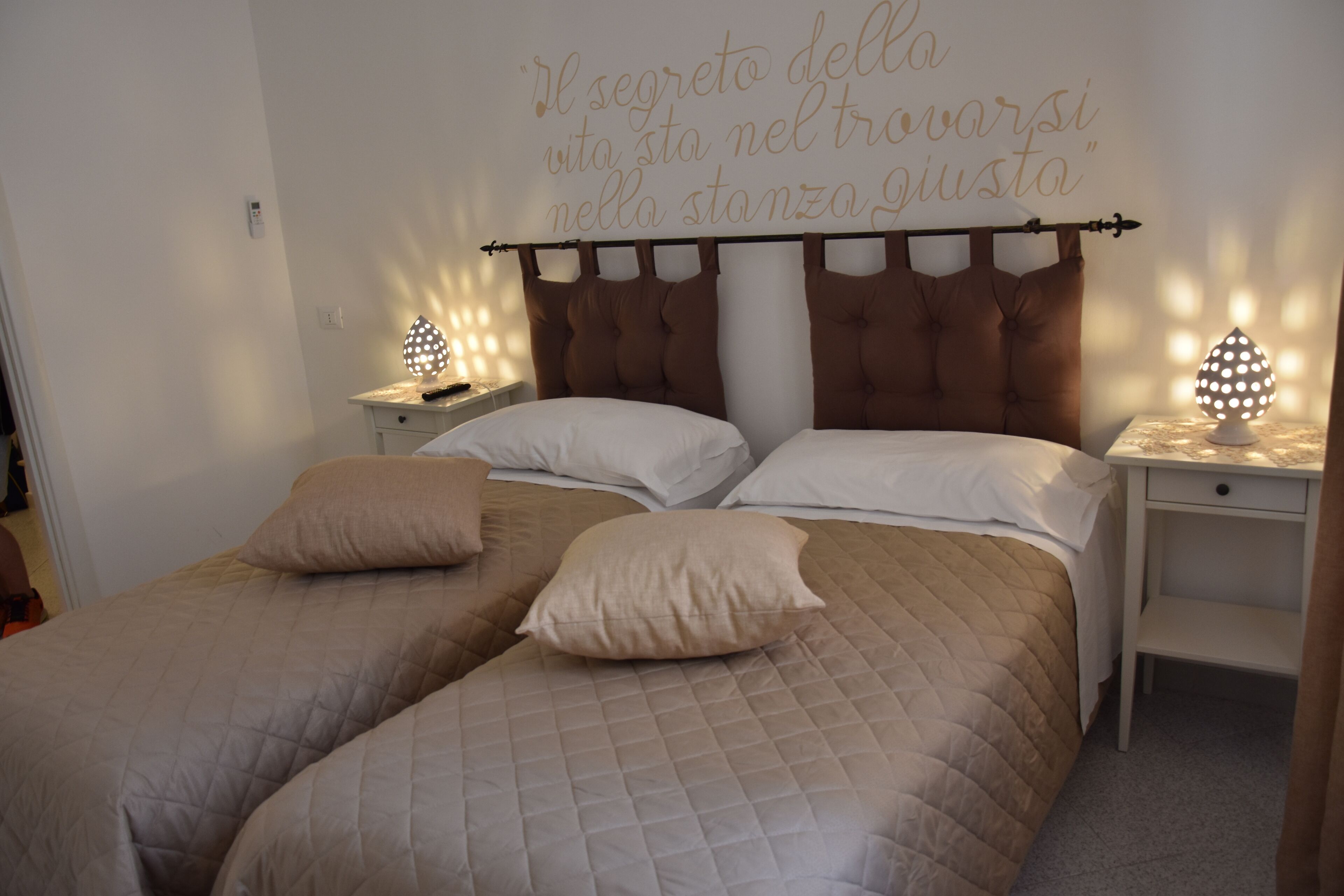 En dobbeltseng med gavl, to sengeborde med lamper, og en væg med et italiensk citat.