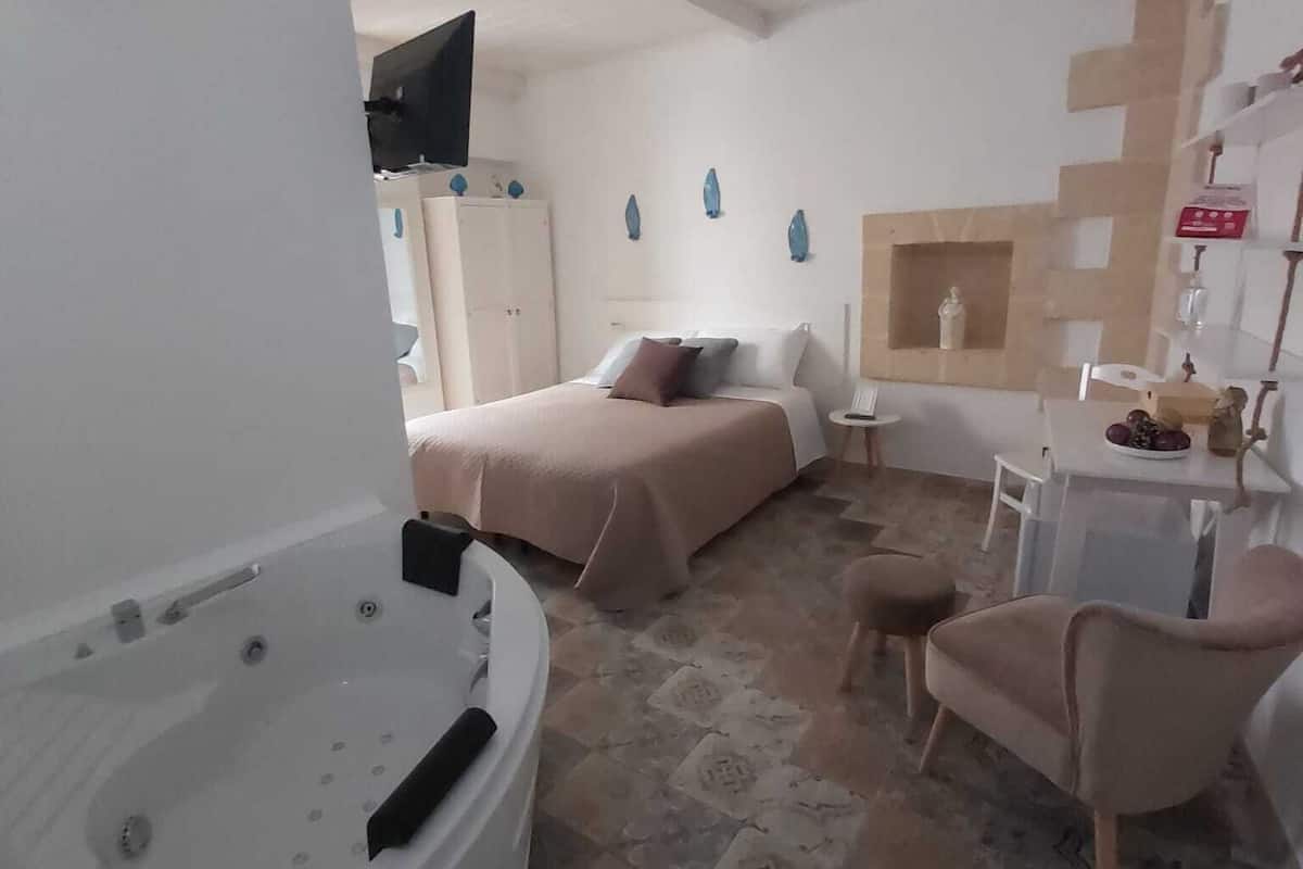 A hotel room with a large bathtub, a bed with two pillows, a small table with a chair, and a TV mounted on the wall.