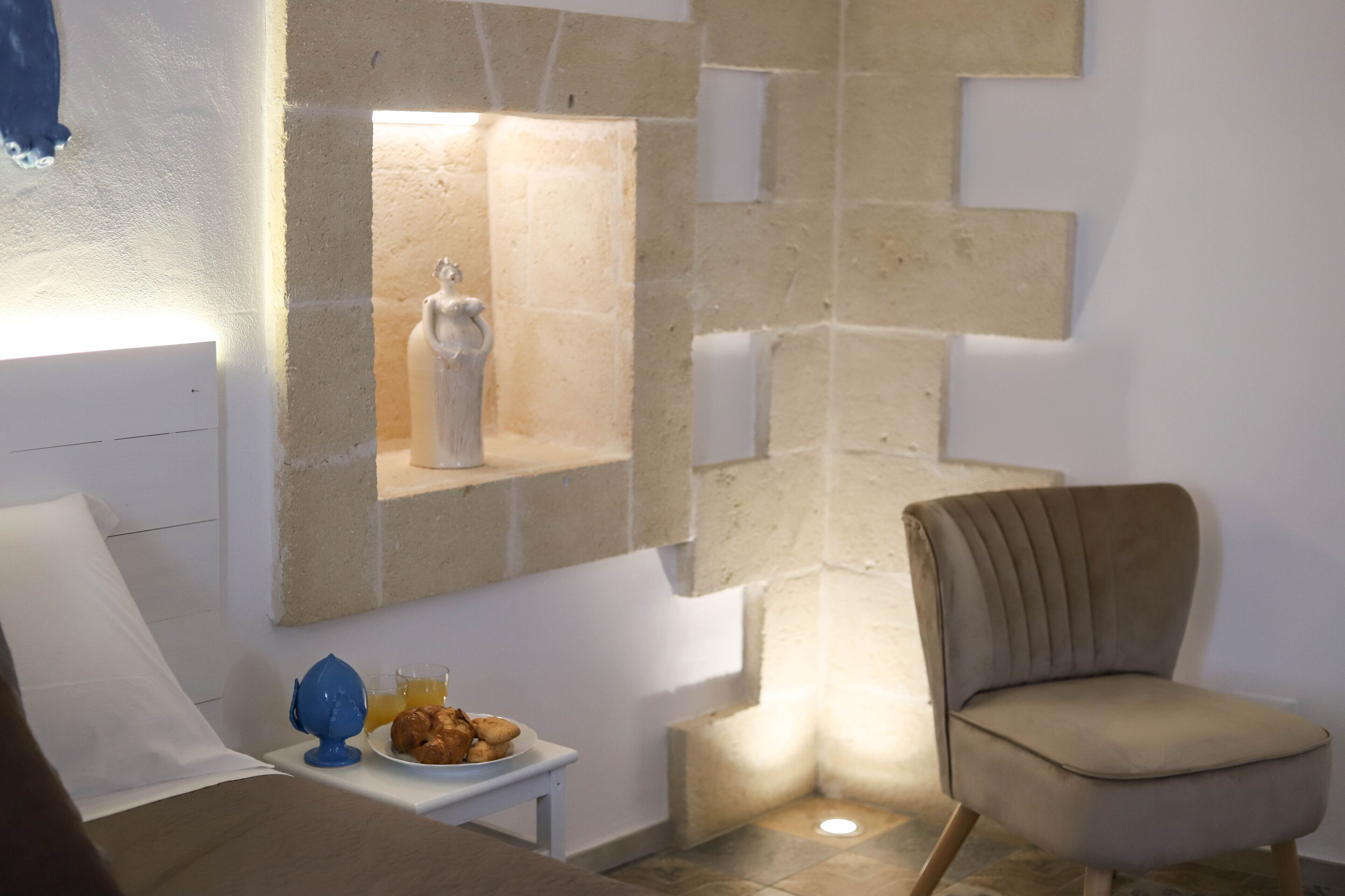Una stanza moderna con una parete in pietra decorativa, una poltrona beige, un tavolino con un piatto di cibo e un oggetto blu, e un cuscino bianco.