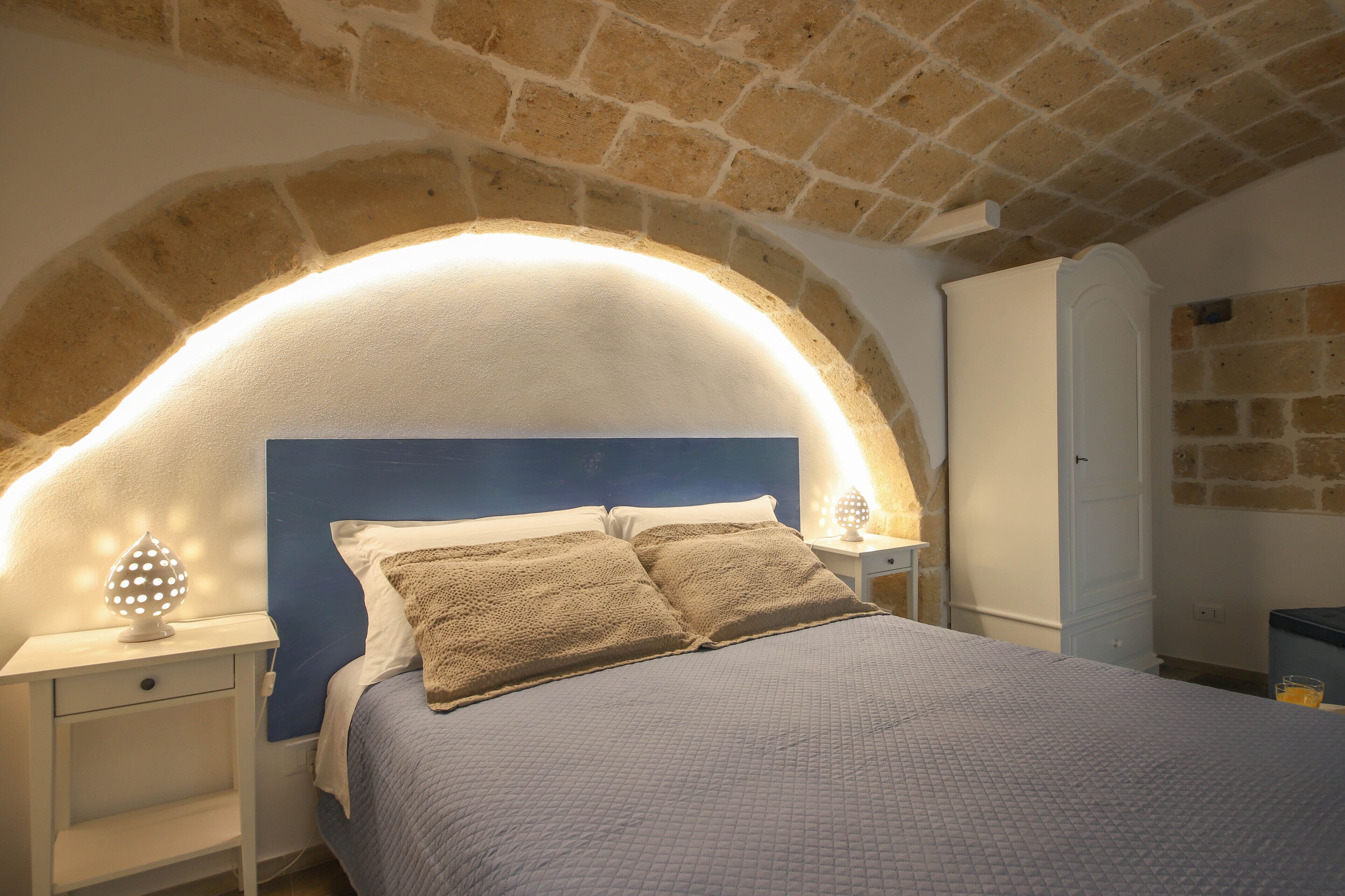 A bedroom with a stone arched ceiling, a bed with a quilted cover, a nightstand with a lamp, and a wardrobe.