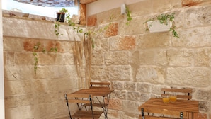 A rustic stone wall with a wooden table and chairs, a hanging plant, and a small shelf with decorative items.